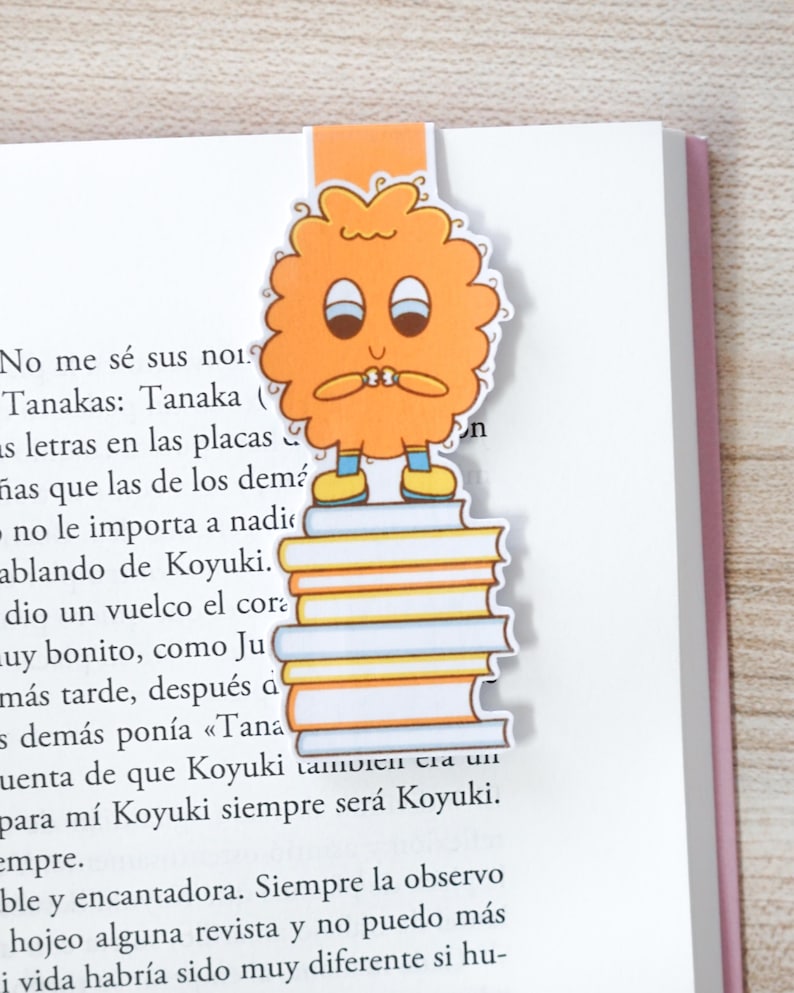 Op de afbeelding: Oranje bladwijzer met een stripfiguur op een stapel boeken, ingevoegd in een boek. Het personage heeft een bezorgde uitdrukking, een rond lichaam, twee ogen en kleine armen. Het boek heeft Spaanse tekst.