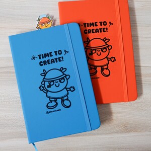 May include: Two notebooks, one blue and one orange, with the text "TIME TO CREATE!" and a cartoon character illustration. A small bookmark with the same character is also visible. The notebooks have elastic closures and ribbon page markers.
