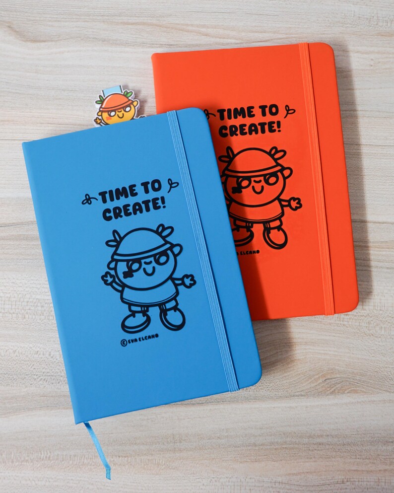 May include: Two notebooks, one blue and one orange, with the text "TIME TO CREATE!" and a cartoon character illustration. A small bookmark with the same character is also visible. The notebooks have elastic closures and ribbon page markers.