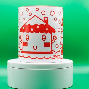 May include: A white ceramic mug with a red cartoon house design. The house has a smiling face and is surrounded by red circles. The mug is on a white platform against a green background.