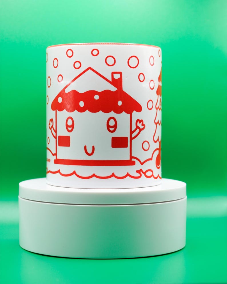 May include: A white ceramic mug with a red cartoon house design. The house has a smiling face and is surrounded by red circles. The mug is on a white platform against a green background.