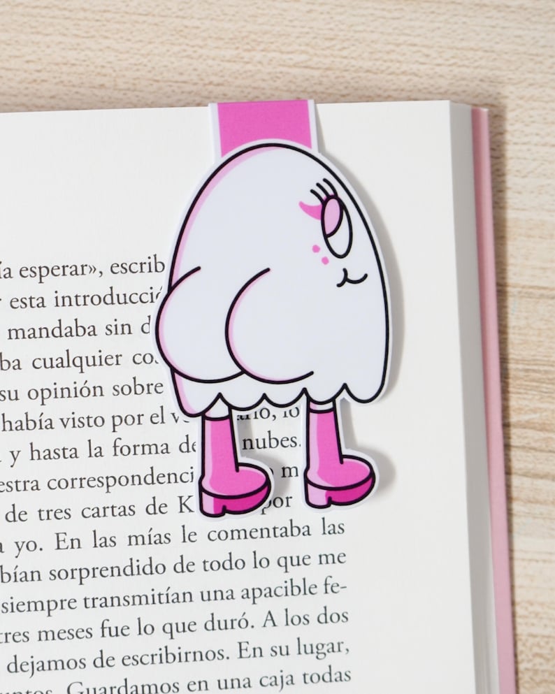 K&ouml;nnte beinhalten: Ein skurriles Lesezeichen mit einem Cartoon-Geist-Design. Der wei&szlig;e Geist hat rosa Stiefel und einen rosa Tab. Das Lesezeichen befindet sich auf den Seiten eines Buches.