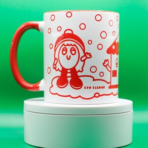 May include: White ceramic mug with a red handle and rim, featuring a whimsical red illustration of a character and a house. The mug has the text "EVA ELCANO" printed on it, and is set against a green background.