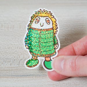 May include: A whimsical sticker featuring a cartoon character with a white face, orange eyes, and yellow spiky hair. The character wears a green coat with glitter accents and green shoes. The sticker is held by a hand.