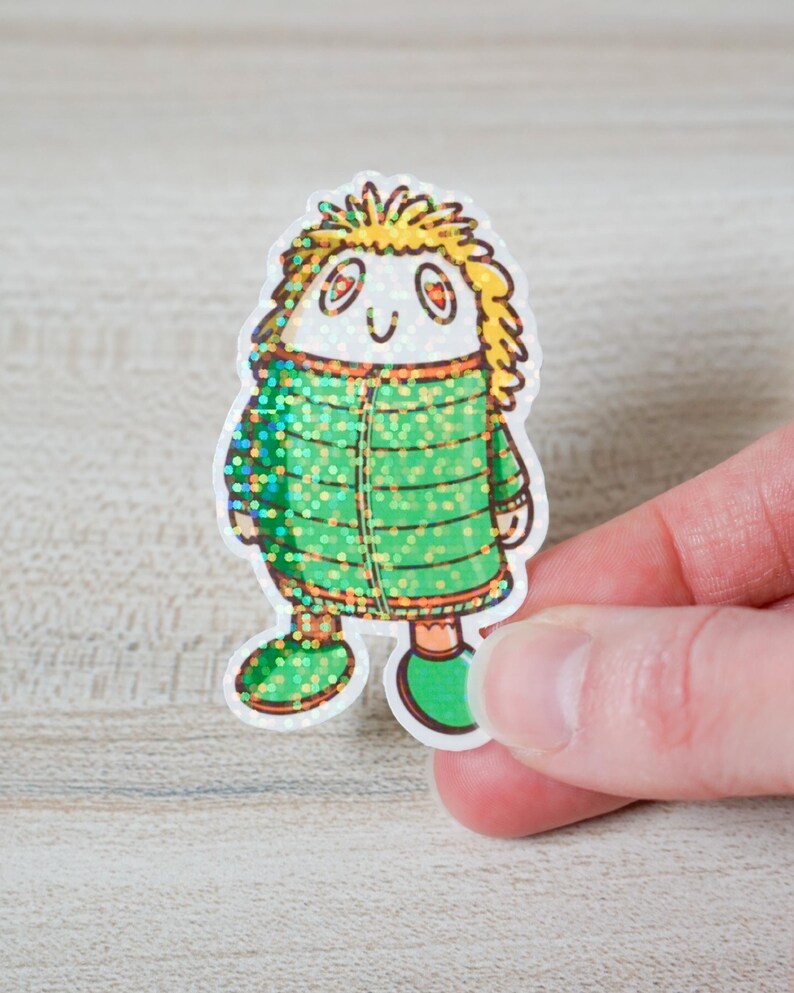 May include: A whimsical sticker featuring a cartoon character with a white face, orange eyes, and yellow spiky hair. The character wears a green coat with glitter accents and green shoes. The sticker is held by a hand.