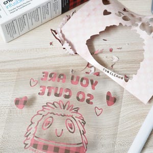 Puede incluir: Una l&aacute;mina de transferencia transparente con el texto "YOU ARE SO CUTE" y un personaje de dibujos animados con una bufanda a cuadros rosa y negro. Una caja de Cricut Infusible Ink y papel recortado en el fondo.