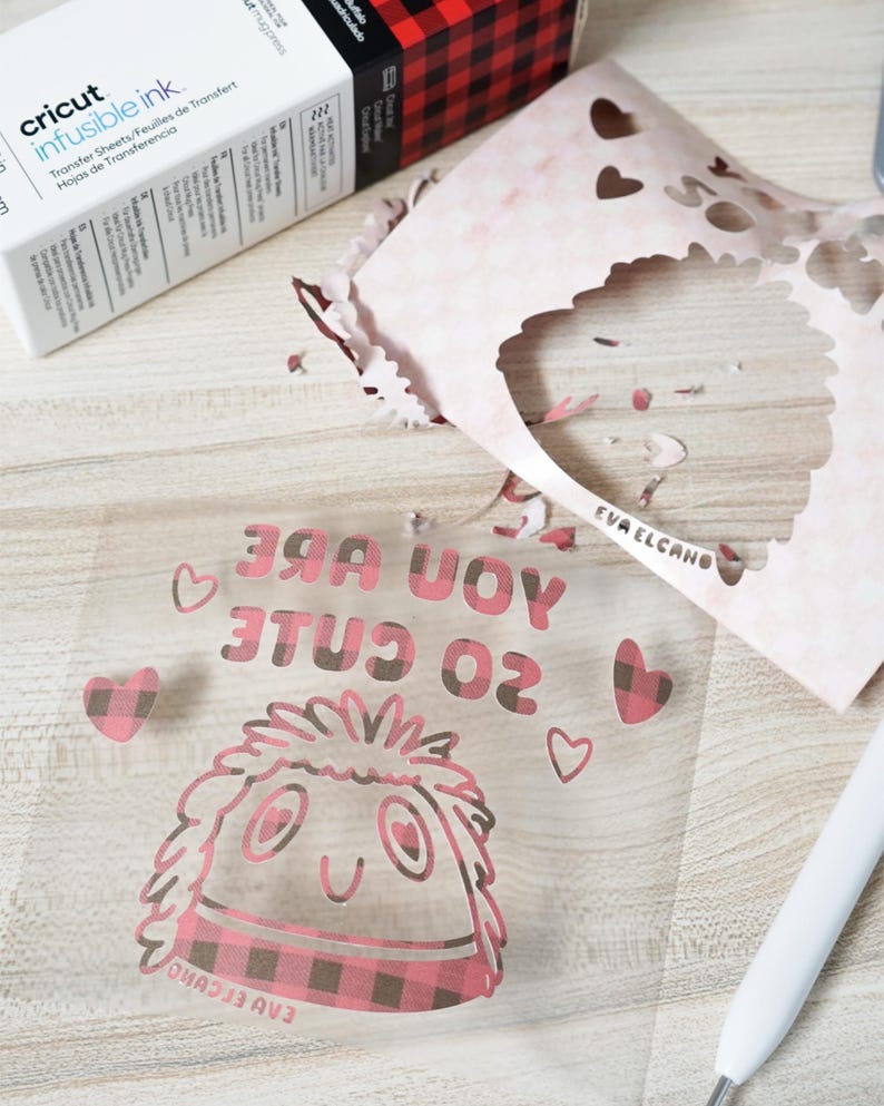 Puede incluir: Una l&aacute;mina de transferencia transparente con el texto "YOU ARE SO CUTE" y un personaje de dibujos animados con una bufanda a cuadros rosa y negro. Una caja de Cricut Infusible Ink y papel recortado en el fondo.