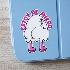 May include: A white cartoon ghost sticker with pink boots and the text "ESTOY DE MIEDO" in a pink circular border. The sticker is on a light blue surface.
