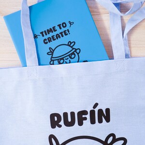 Może przedstawiać: Jasnoniebieska torba z nadrukiem "RUFIN" w kolorze czarnym. Wewnątrz torby znajduje się niebieski notatnik z napisem "TIME TO CREATE!" i postacią z kresk&oacute;wki. Torba ma jasnoniebieskie paski.