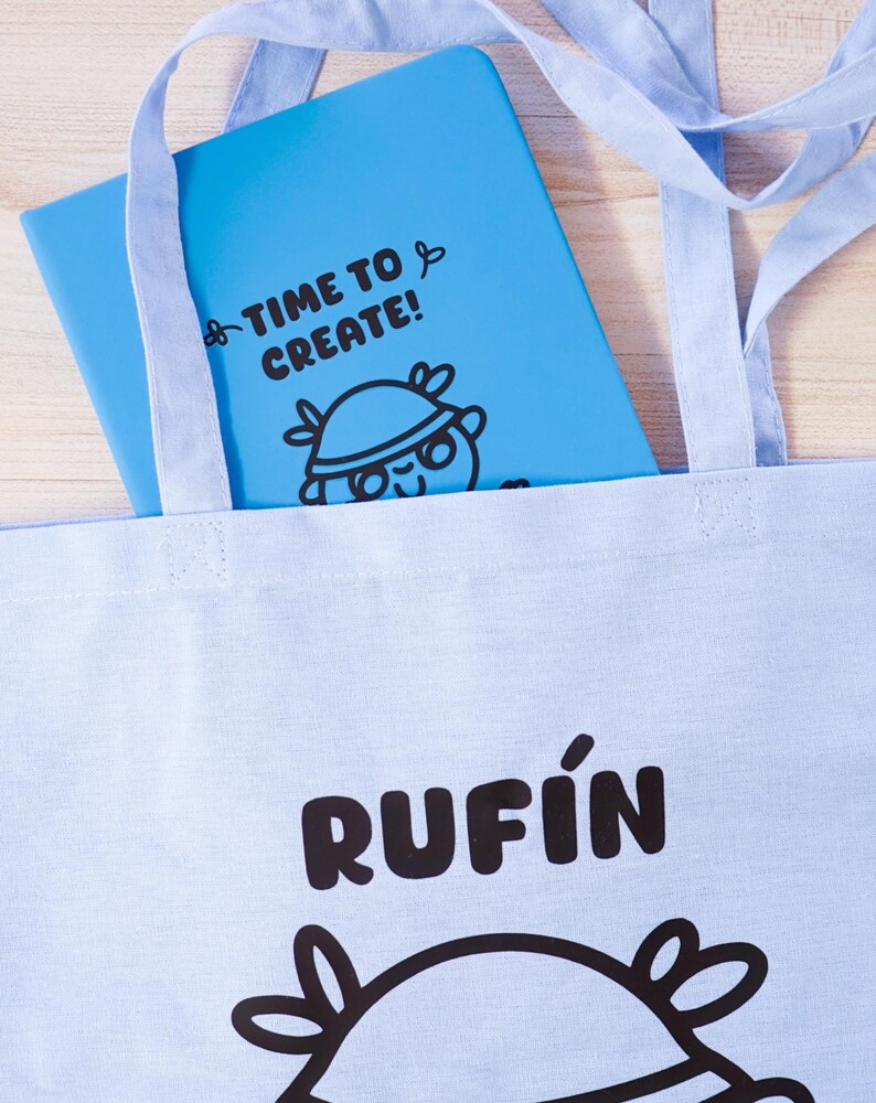 Może przedstawiać: Jasnoniebieska torba z nadrukiem "RUFIN" w kolorze czarnym. Wewnątrz torby znajduje się niebieski notatnik z napisem "TIME TO CREATE!" i postacią z kresk&oacute;wki. Torba ma jasnoniebieskie paski.