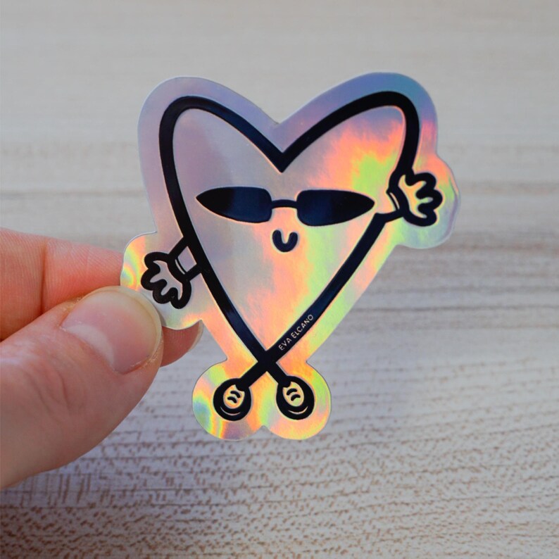 Puede incluir: Pegatina hologr&aacute;fica con un personaje en forma de coraz&oacute;n con gafas de sol y brazos extendidos. La pegatina tiene un contorno negro y el texto "EVA ELCANO". La pegatina est&aacute; sujeta por los dedos de una persona.