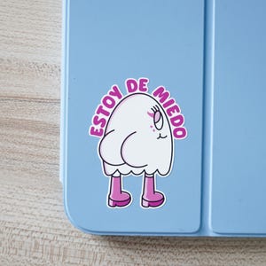May include: A white cartoon ghost sticker with pink accents and the Spanish text "ESTOY DE MIEDO" on a light blue surface. The ghost has pink feet and a worried expression. The sticker is likely for a laptop or water bottle.