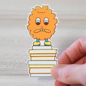 Puede incluir: Una pegatina con un personaje de dibujos animados con un cuerpo naranja y esponjoso, de pie sobre una pila de libros. El personaje tiene una expresión preocupada, con las manos juntas cerca de la boca. Los libros son blancos, amarillos y naranjas.