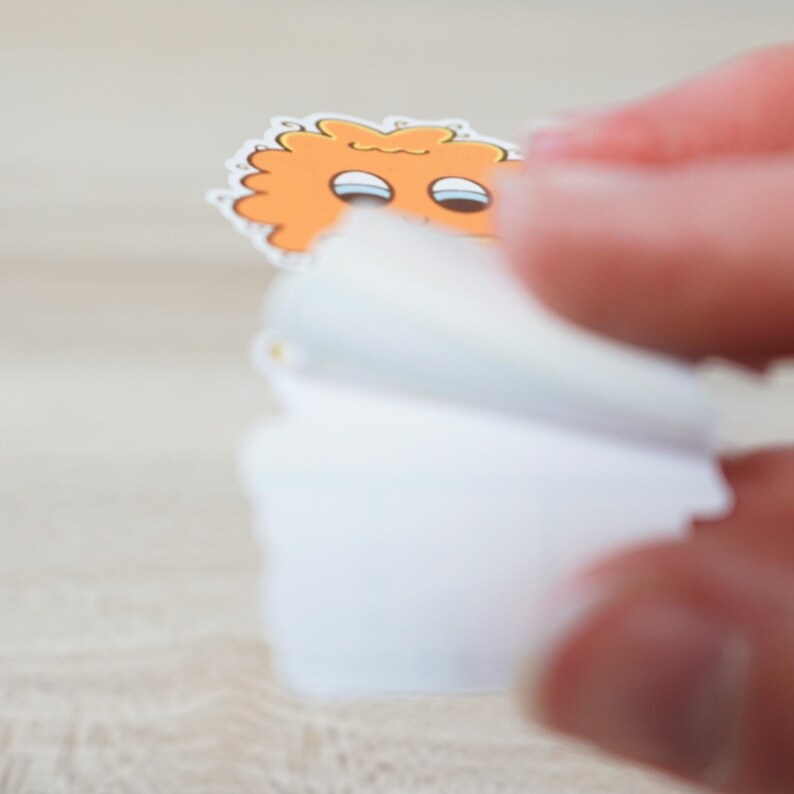 May include: A small, orange sticker with a cartoon monster design is being peeled from a stack of white stickers. The monster has two eyes and a fluffy, textured appearance. The sticker is on a light-colored surface.