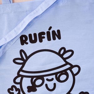 Może przedstawiać: Jasnoniebieska torba typu tote z czarnym napisem "RUFIN". Poniżej znajduje się postać z kresk&oacute;wki w kapeluszu i okularach. Torba ma dwa uchwyty i jest wykonana z tkaniny.