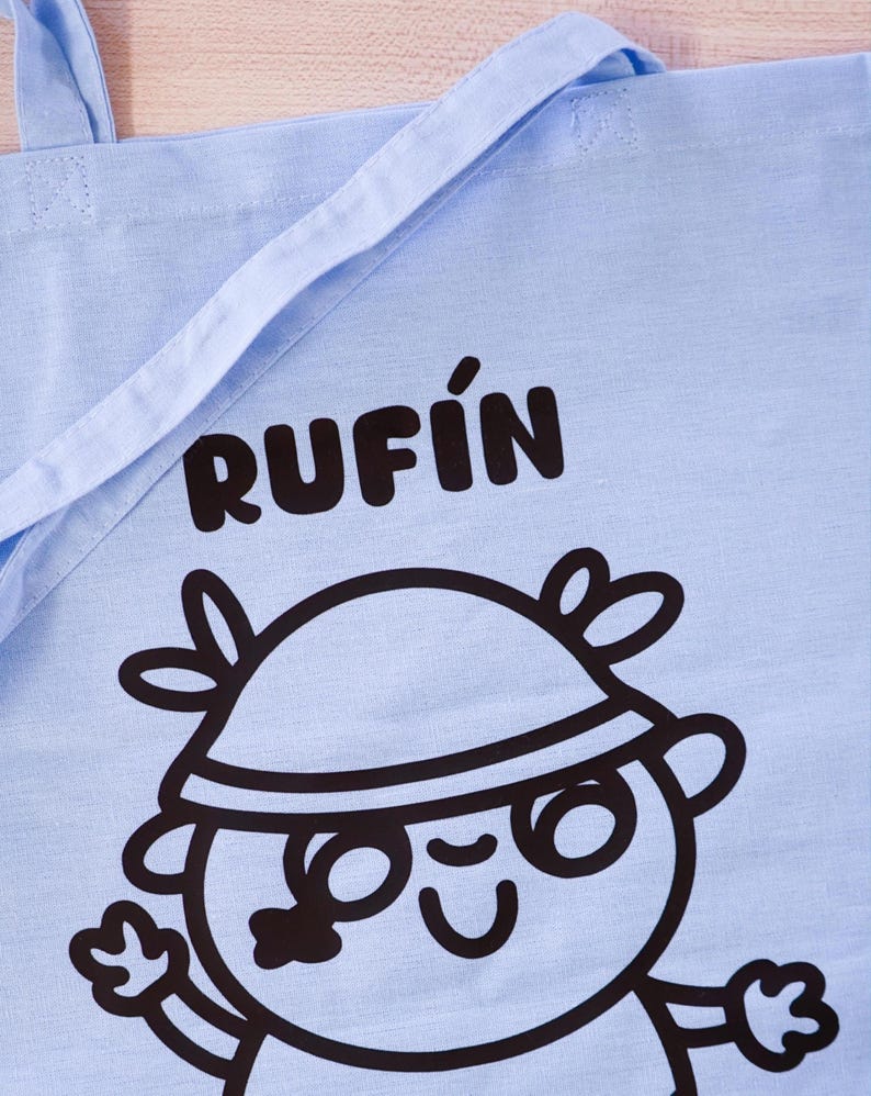 Może przedstawiać: Jasnoniebieska torba typu tote z czarnym napisem "RUFIN". Poniżej znajduje się postać z kresk&oacute;wki w kapeluszu i okularach. Torba ma dwa uchwyty i jest wykonana z tkaniny.
