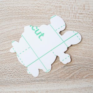 May include: A white turtle-shaped sticker with a green grid pattern and the word "cut" printed on it. The sticker has a protective layer and is designed for crafting projects. The sticker is on a light brown wooden surface.