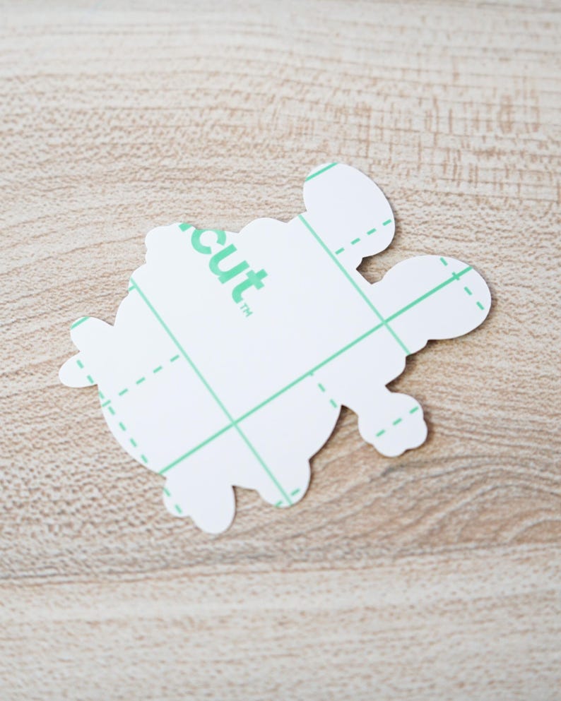 May include: A white turtle-shaped sticker with a green grid pattern and the word "cut" printed on it. The sticker has a protective layer and is designed for crafting projects. The sticker is on a light brown wooden surface.