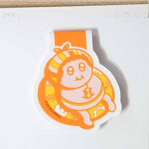 May include: An orange and yellow baby-themed bookmark with a cartoon illustration. The bookmark features a smiling baby in a yellow and orange striped hat and a bib with a bell. The text "EVA ELCANO" is printed on the bookmark.