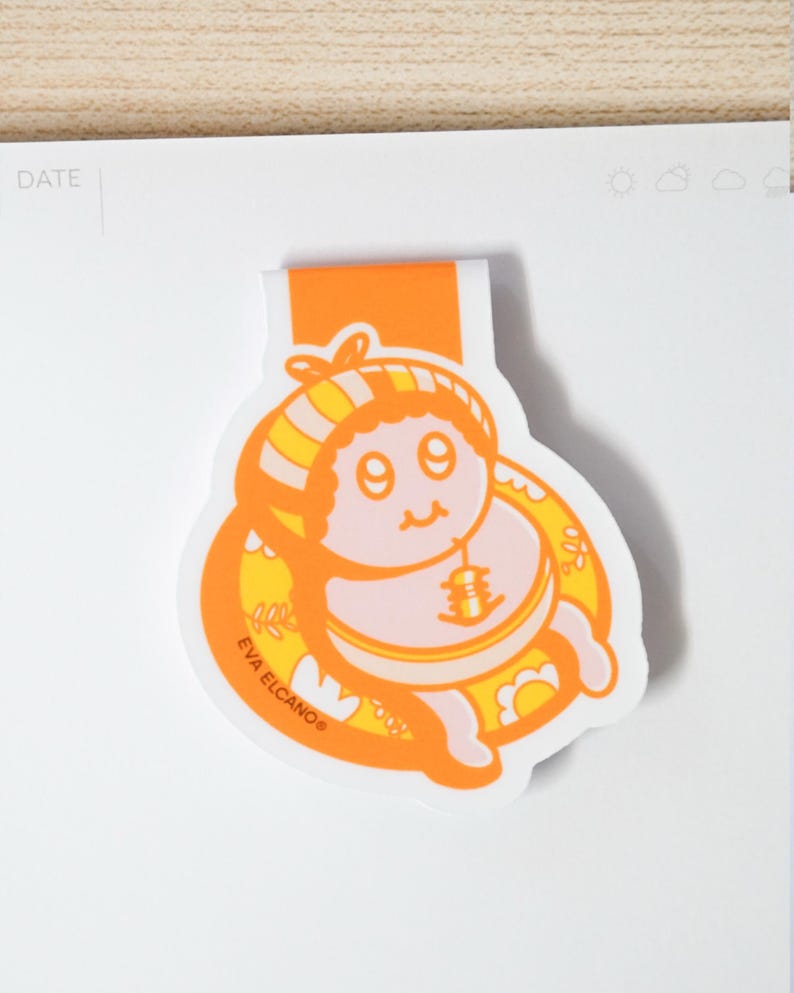 May include: An orange and yellow baby-themed bookmark with a cartoon illustration. The bookmark features a smiling baby in a yellow and orange striped hat and a bib with a bell. The text "EVA ELCANO" is printed on the bookmark.