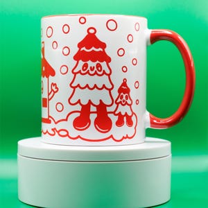 May include: White ceramic mug with a red interior and handle. The mug features a whimsical red illustration of a Christmas tree and a smaller tree, with red circles resembling snowflakes. The mug is set against a vibrant green background.