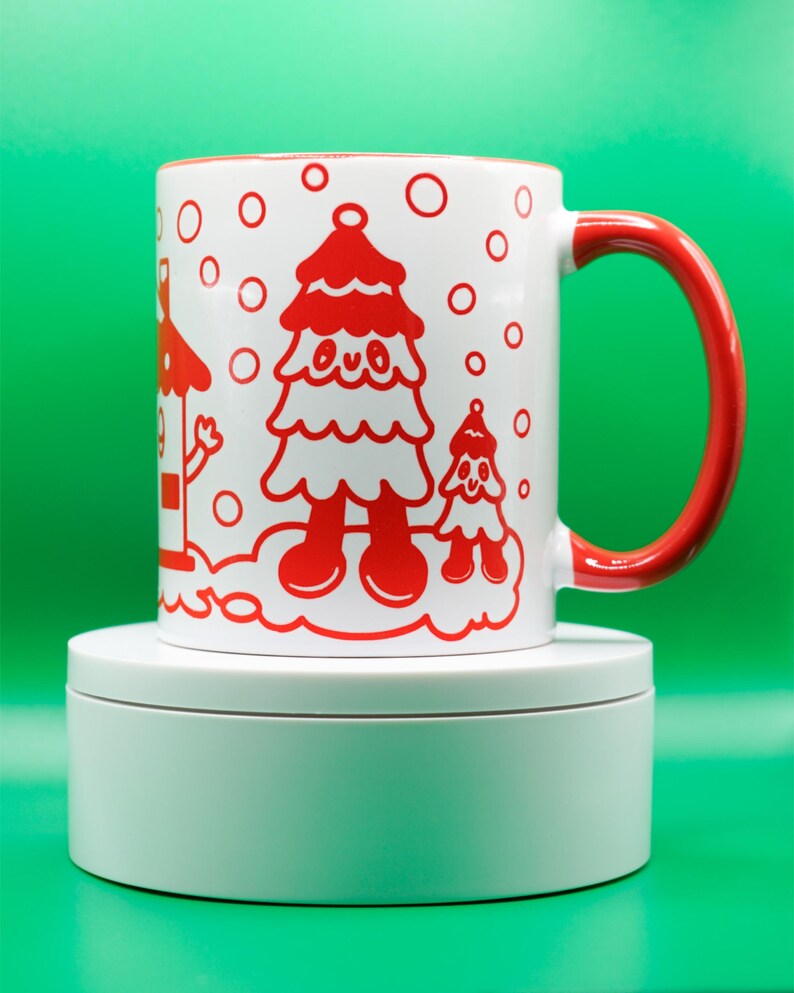 May include: White ceramic mug with a red interior and handle. The mug features a whimsical red illustration of a Christmas tree and a smaller tree, with red circles resembling snowflakes. The mug is set against a vibrant green background.