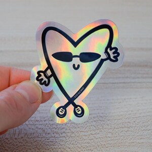 Puede incluir: Pegatina hologr&aacute;fica con un personaje en forma de coraz&oacute;n con gafas de sol, brazos y piernas. El personaje tiene una cara sonriente y est&aacute; delineado en negro. La pegatina tiene un brillo arco&iacute;ris y el texto "EVA ELCANO".