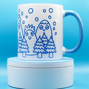 May include: White ceramic mug with a blue handle and a whimsical mountain and tree design in blue. The design features smiling mountains, trees, and falling snow. The mug is sitting on a light blue platform against a blue background.