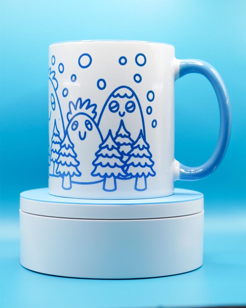 May include: White ceramic mug with a blue handle and a whimsical mountain and tree design in blue. The design features smiling mountains, trees, and falling snow. The mug is sitting on a light blue platform against a blue background.