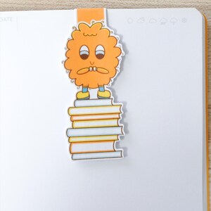 Op de afbeelding: Oranje bladwijzer met een stripfiguur op een stapel boeken. Het personage heeft een bezorgde uitdrukking. De bladwijzer is bevestigd aan een wit notitieboek met een oranje rand.