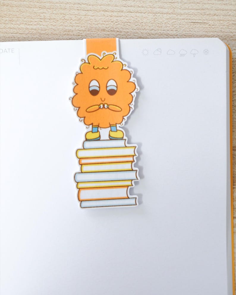 Op de afbeelding: Oranje bladwijzer met een stripfiguur op een stapel boeken. Het personage heeft een bezorgde uitdrukking. De bladwijzer is bevestigd aan een wit notitieboek met een oranje rand.