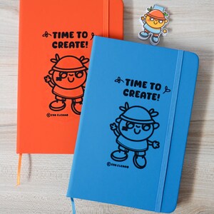 May include: Two notebooks, one orange and one blue, featuring the text "TIME TO CREATE!" and a cartoon character illustration. A small sticker of the same character is also present. The notebooks have a ribbon bookmark and the brand name "EVA ELCANO" is printed.