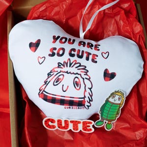 Puede incluir: Una almohada blanca en forma de coraz&oacute;n con el texto "YOU ARE SO CUTE" en rojo y negro. La almohada presenta un personaje de dibujos animados y peque&ntilde;os corazones. Tambi&eacute;n hay una pegatina "CUTE" y una pegatina de personaje peque&ntilde;o.