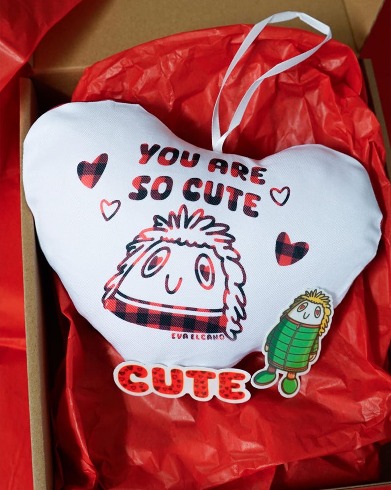 Puede incluir: Una almohada blanca en forma de coraz&oacute;n con el texto "YOU ARE SO CUTE" en rojo y negro. La almohada presenta un personaje de dibujos animados y peque&ntilde;os corazones. Tambi&eacute;n hay una pegatina "CUTE" y una pegatina de personaje peque&ntilde;o.