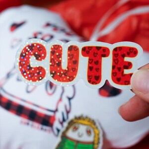Puede incluir: Una pegatina brillante con la palabra "CUTE" en fuente de burbujas. Las letras son rojas con patrones de coraz&oacute;n y detalles de purpurina. Un peque&ntilde;o personaje de dibujos animados y un objeto blanco con un patr&oacute;n de cuadros rojos y negros est&aacute;n en el fondo.