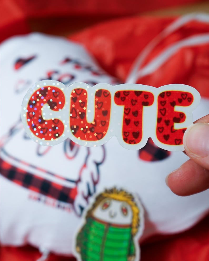 Puede incluir: Una pegatina brillante con la palabra "CUTE" en fuente de burbujas. Las letras son rojas con patrones de coraz&oacute;n y detalles de purpurina. Un peque&ntilde;o personaje de dibujos animados y un objeto blanco con un patr&oacute;n de cuadros rojos y negros est&aacute;n en el fondo.