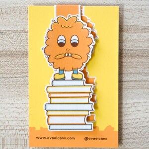 Op de afbeelding: Een grillige boekenlegger met een stripfiguur met oranje vacht die op een stapel boeken staat. De boekenlegger is geel en oranje met de tekst "www.evaelcano.com" en "@evaelcano". Het personage heeft een bezorgde uitdrukking.