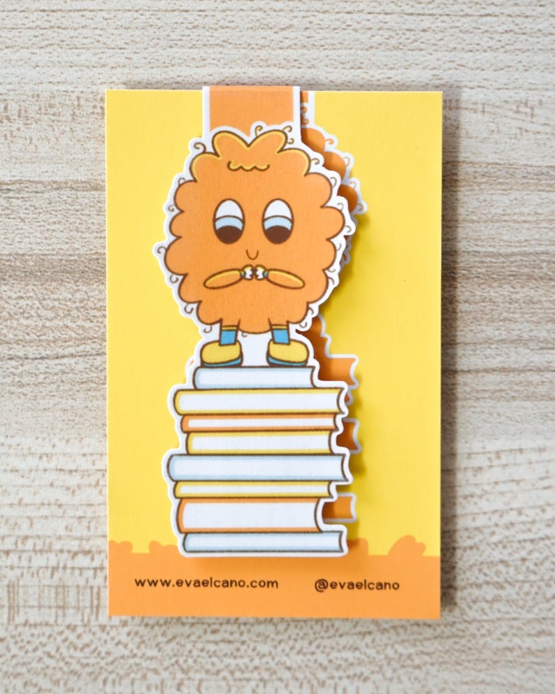 Op de afbeelding: Een grillige boekenlegger met een stripfiguur met oranje vacht die op een stapel boeken staat. De boekenlegger is geel en oranje met de tekst "www.evaelcano.com" en "@evaelcano". Het personage heeft een bezorgde uitdrukking.