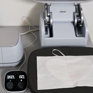 Puede incluir: Una m&aacute;quina de prensa de calor Cricut con un panel de control digital que muestra 350 grados y 60 segundos. Una hoja de material blanco descansa sobre una superficie de prensa de calor negra y redondeada. La m&aacute;quina es gris claro.