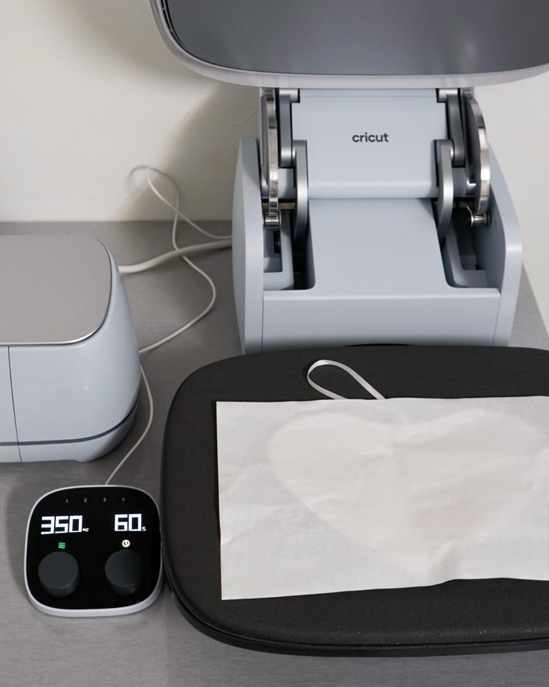 Puede incluir: Una m&aacute;quina de prensa de calor Cricut con un panel de control digital que muestra 350 grados y 60 segundos. Una hoja de material blanco descansa sobre una superficie de prensa de calor negra y redondeada. La m&aacute;quina es gris claro.