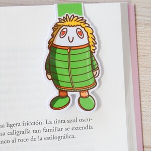 May include: A whimsical bookmark featuring a cartoon character with yellow hair, heart-shaped eyes, and a green jacket. The bookmark is inserted into a book, with the character's head and a green tab visible above the pages.