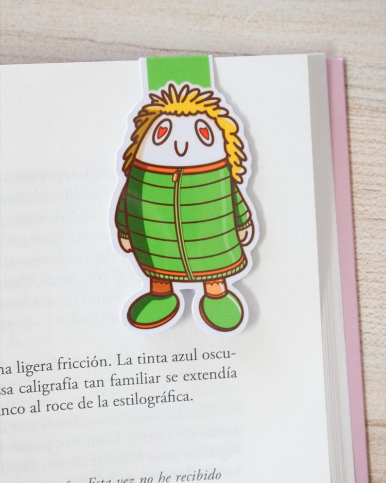 May include: A whimsical bookmark featuring a cartoon character with yellow hair, heart-shaped eyes, and a green jacket. The bookmark is inserted into a book, with the character's head and a green tab visible above the pages.