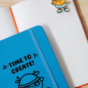 May include: An open notebook with white pages and an orange spine. A blue notebook with the text "TIME TO CREATE!" and a cartoon illustration is in the foreground. A cartoon character bookmark is attached to the notebook pages.