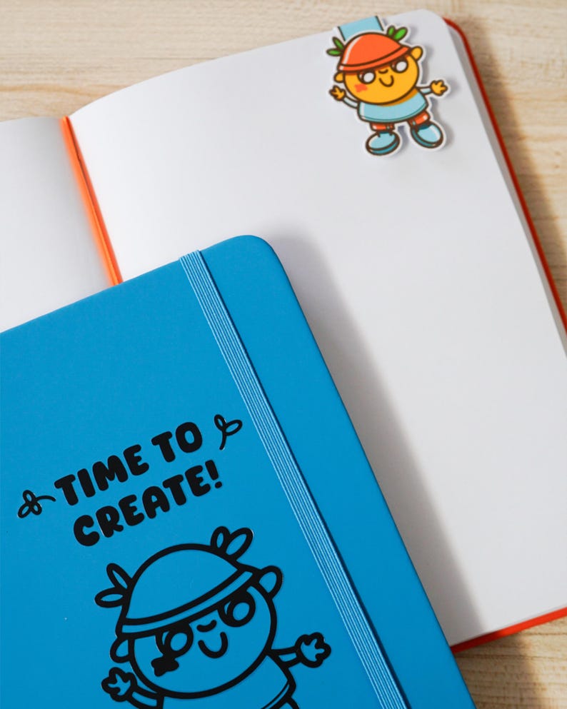 May include: An open notebook with white pages and an orange spine. A blue notebook with the text "TIME TO CREATE!" and a cartoon illustration is in the foreground. A cartoon character bookmark is attached to the notebook pages.