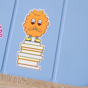 May include: A light blue surface with a cartoon sticker of an orange, fluffy character with big eyes and small shoes, standing on a stack of books. The books have white, yellow, and orange pages.