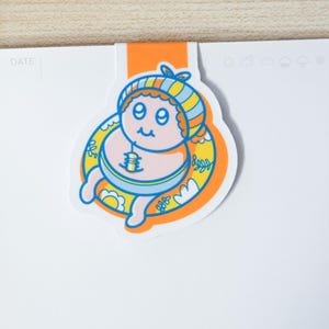 May include: A colorful cartoon bookmark featuring a smiling character in a striped hat and a yellow floatie. The bookmark is orange and white, with the word "DATE" visible on the attached page. The character has a blue and yellow striped hat.