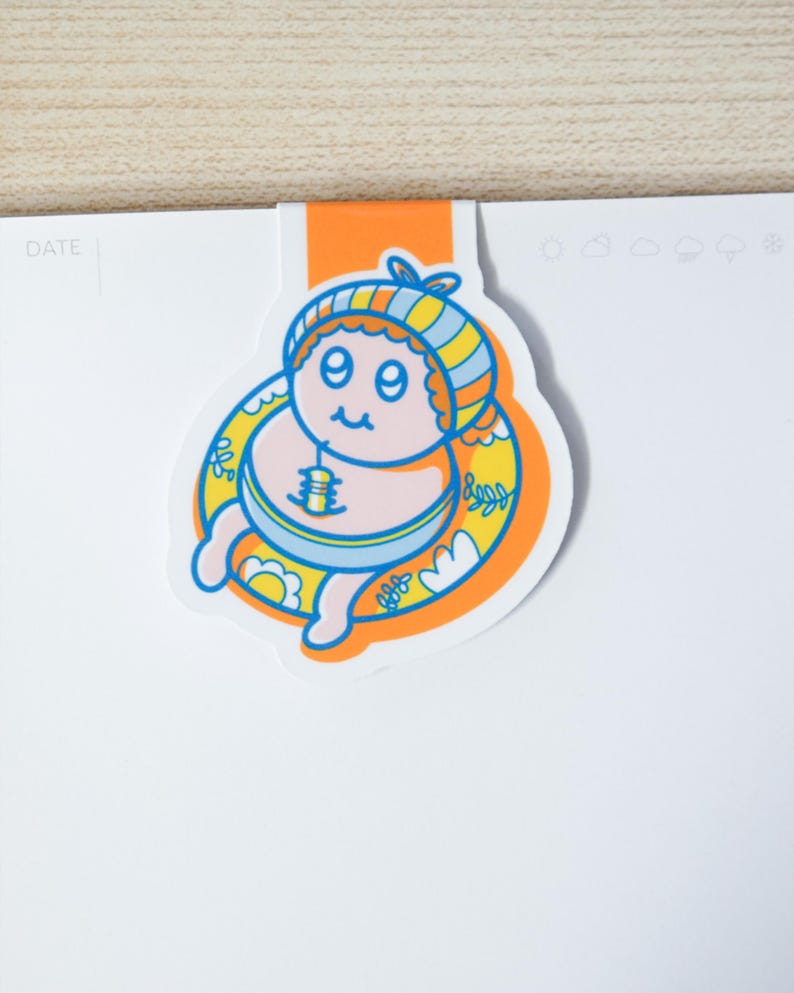 May include: A colorful cartoon bookmark featuring a smiling character in a striped hat and a yellow floatie. The bookmark is orange and white, with the word "DATE" visible on the attached page. The character has a blue and yellow striped hat.