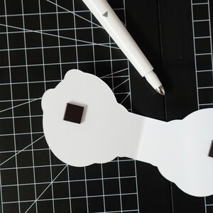 May include: A white pen with a gray triangle detail rests on a black cutting mat with a grid pattern. A white, cloud-shaped object with a small, dark square magnet is also on the mat.
