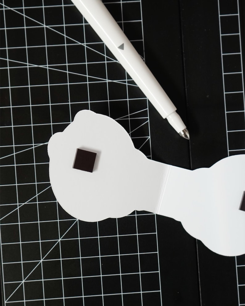 May include: A white pen with a gray triangle detail rests on a black cutting mat with a grid pattern. A white, cloud-shaped object with a small, dark square magnet is also on the mat.