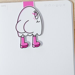 K&ouml;nnte beinhalten: Ein wei&szlig;er, cartoonartiger Lesezeichen in Geisterform mit rosa Stiefeln und einem rosa Oberteil. Das Lesezeichen ist an einem wei&szlig;en Notizbuch befestigt. Der Geist hat ein l&auml;chelndes Gesicht mit rosa Akzenten. Das Notizbuch hat das Wort "DATE" links oben aufgedruckt.